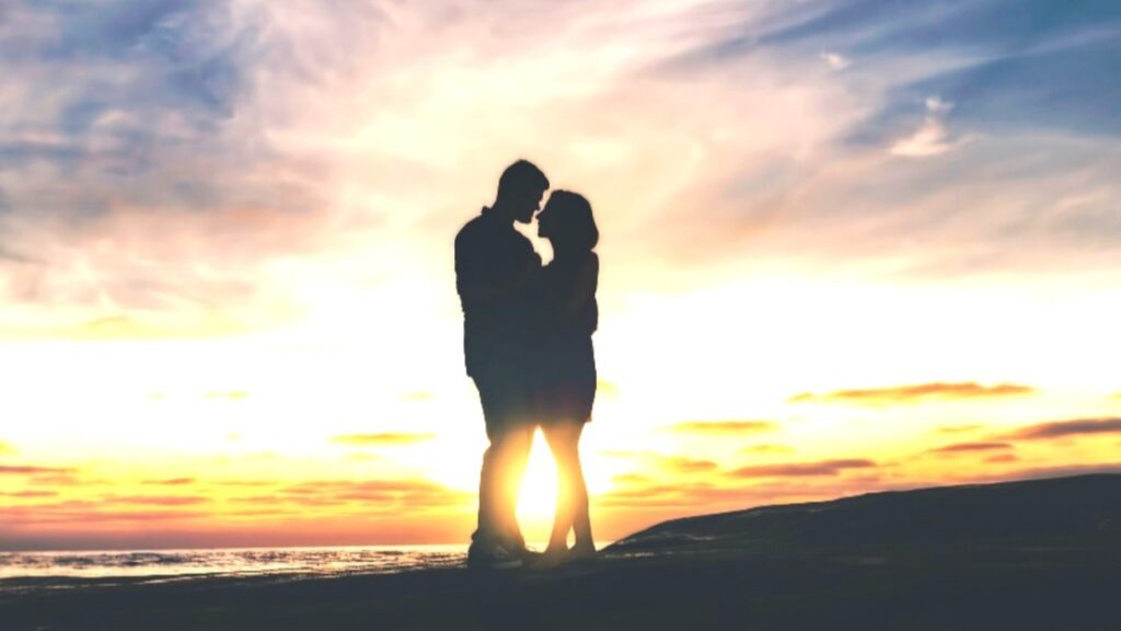 A twin flame reunion depicted by the silhouette of a man and a woman hugging and gazing at each other on a beach at sunset